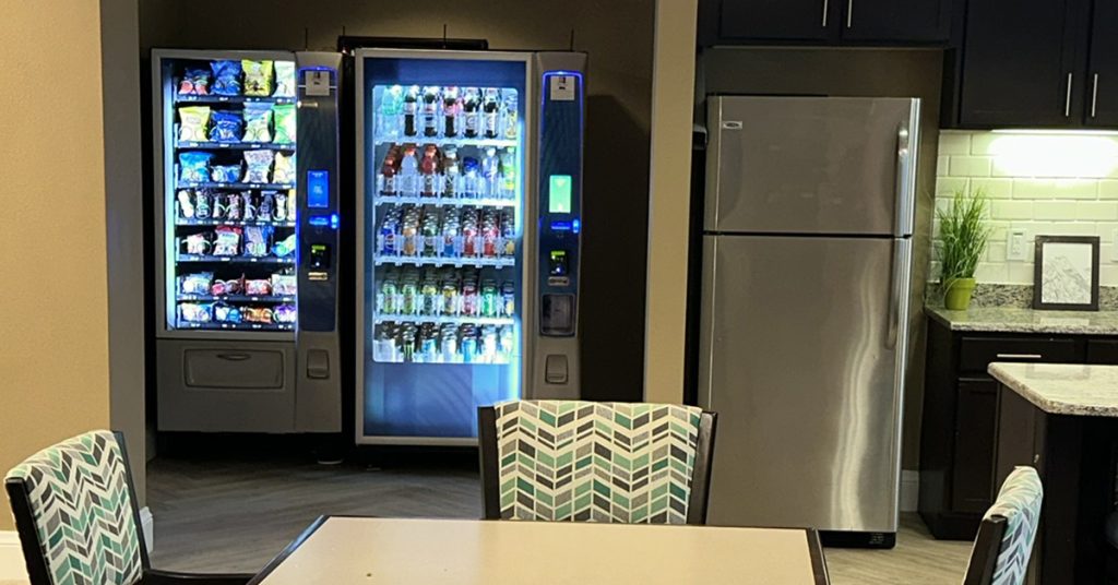 Two vending machines in an office break room, one stocked with snacks and the other with drinks.