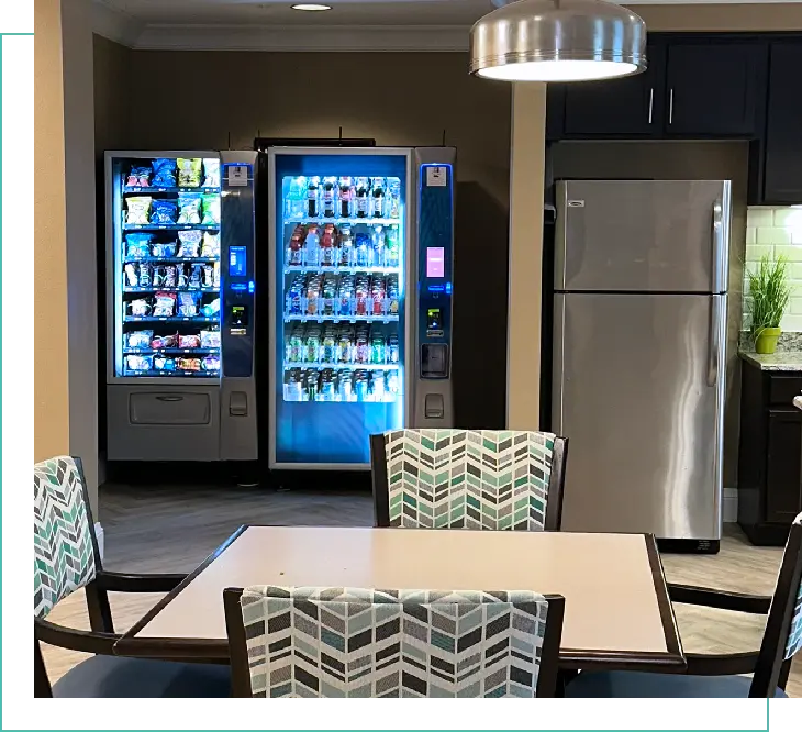 Two vending machines in an office break room, one stocked with snacks and the other with drinks.